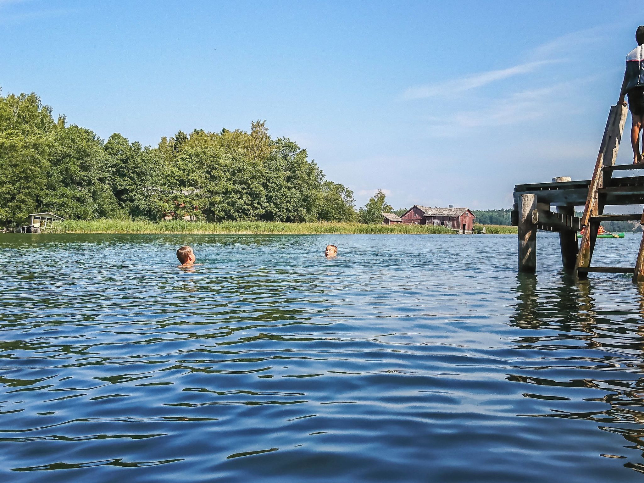 A calm lake with a wooden dock extending into the water, surrounded by lush greenery and a few buildings in the distance.