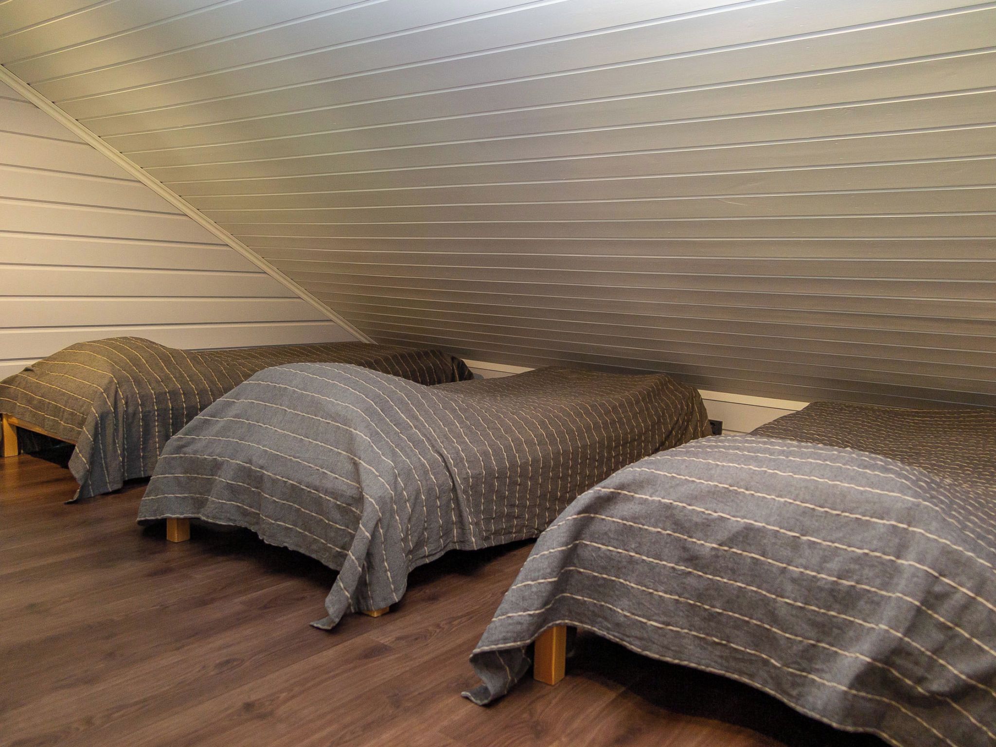 A room with three beds aligned against a sloped ceiling, wooden flooring, and a white wall.
