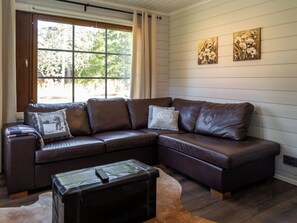 Un sofá modular con varios cojines, un control remoto sobre un reposapiés de cuero y una ventana con cortinas.