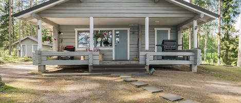 Une cabane avec une véranda, un barbecue et un petit abri de jardin, située dans une zone boisée.