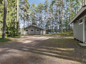 A cabin with a gravel driveway, surrounded by tall trees, and a small dock extending into a body of water.