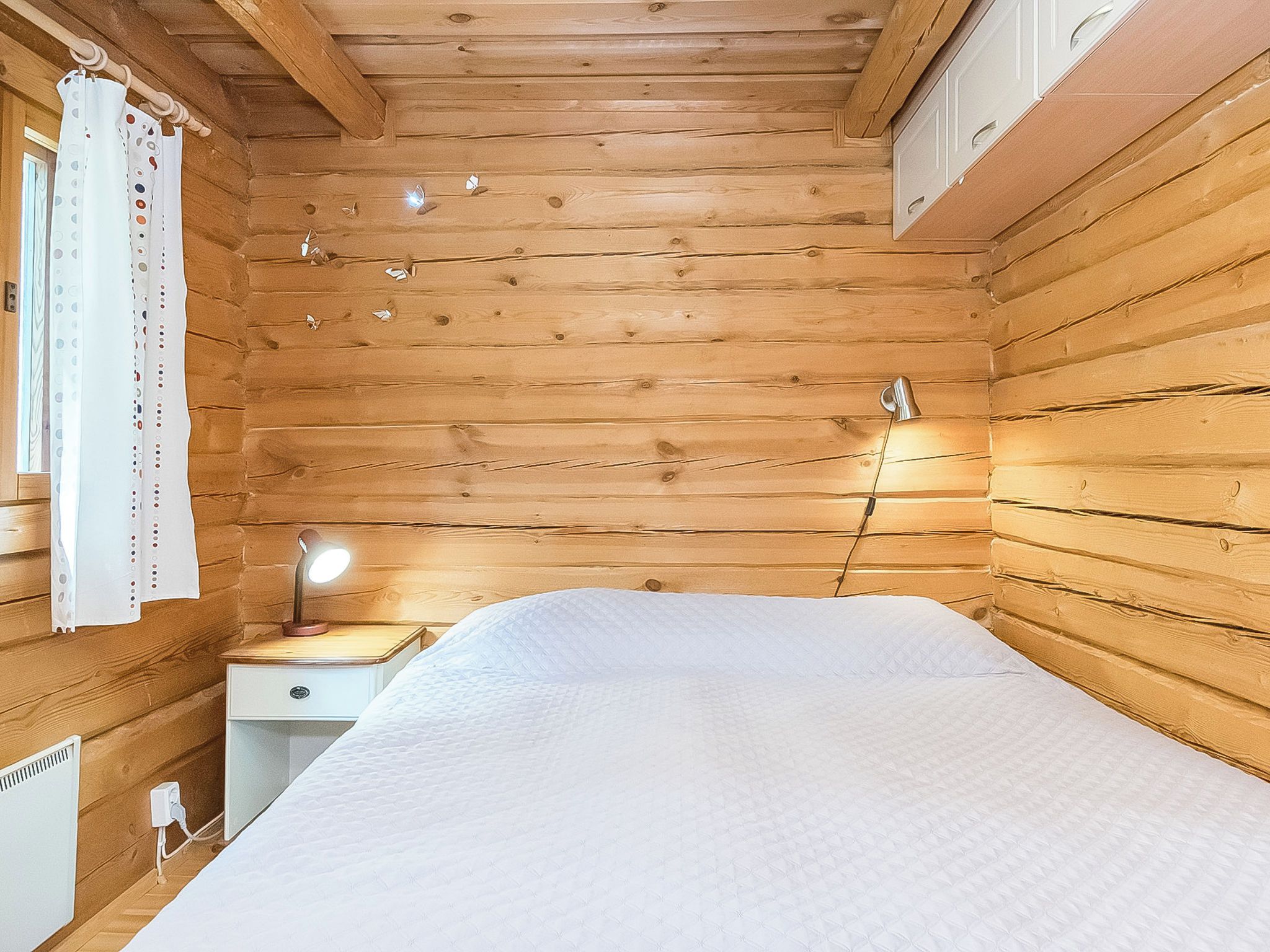 Een houten kamer met een bed, een nachtkastje met een lamp en een raam met gordijnen.