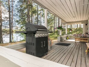 Une terrasse extérieure couverte, équipée d’un barbecue, d’une cheminée en pierre et offrant une vue sur un lac entouré d’arbres.