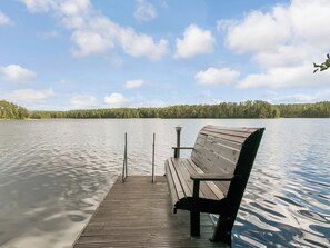 Un muelle de madera con un banco se extiende sobre un lago tranquilo, rodeado de una orilla arbolada.
