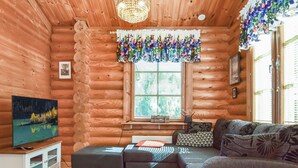 A cozy log cabin interior with a wooden exterior, a large window with blinds, a television on a white cabinet, and a sofa with patterned cushions. - Petäjäniemi by Interhome (Kinnula)