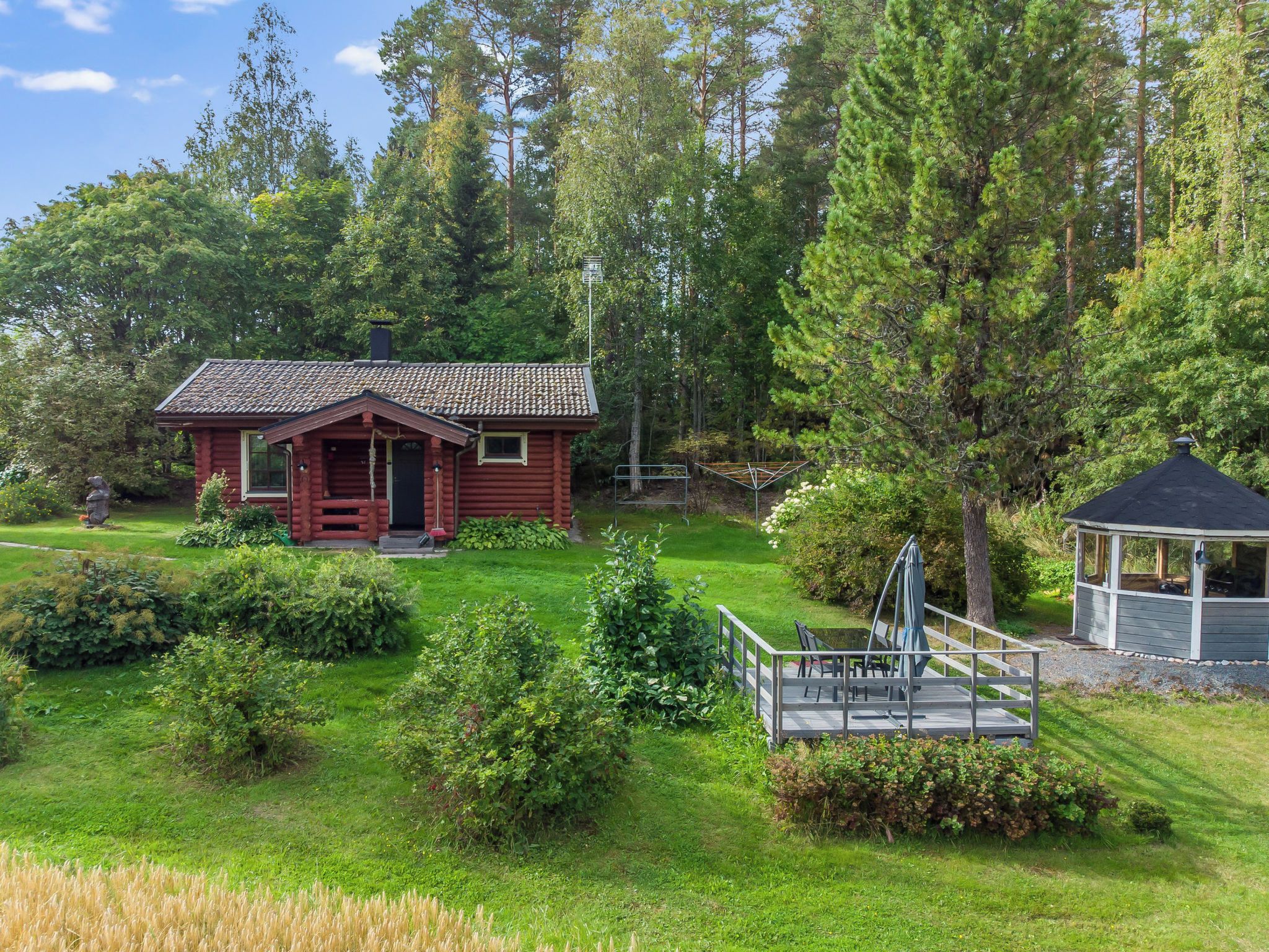 En röd timmerstuga med ett tak täckt av tegelpannor, omgiven av en välskött gräsmatta och en tät skog.
