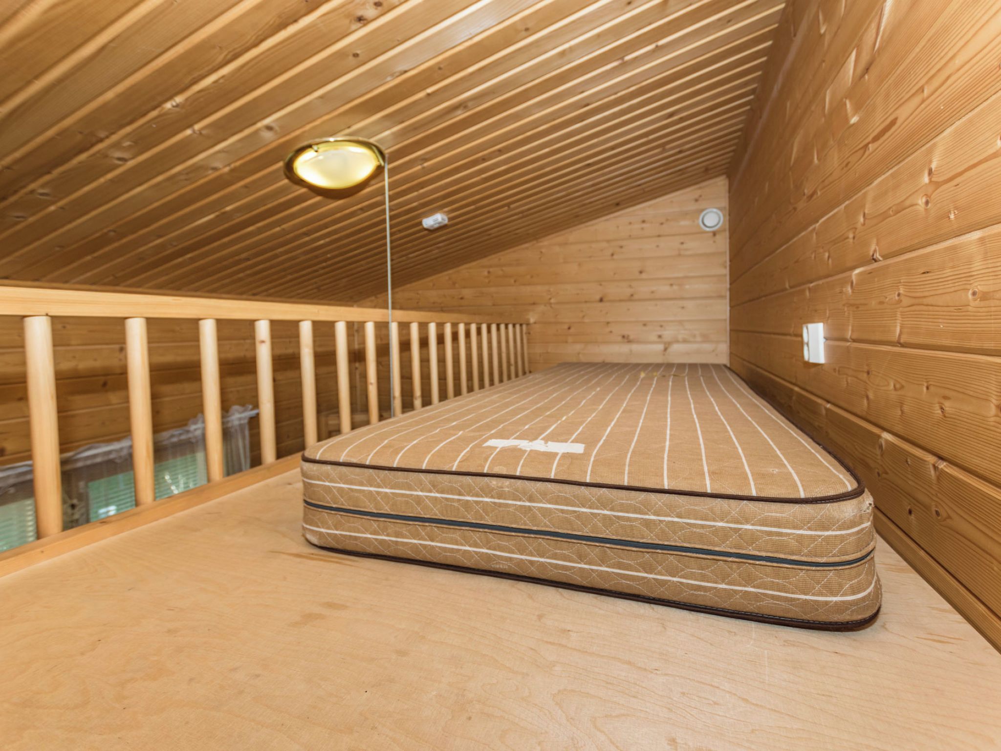 Una habitación de madera con un colchón en el suelo, una barandilla y una lámpara.