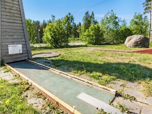 A dilapidated outdoor area with a concrete floor, a wooden structure with a sign, and a grassy field with trees and a large rock.