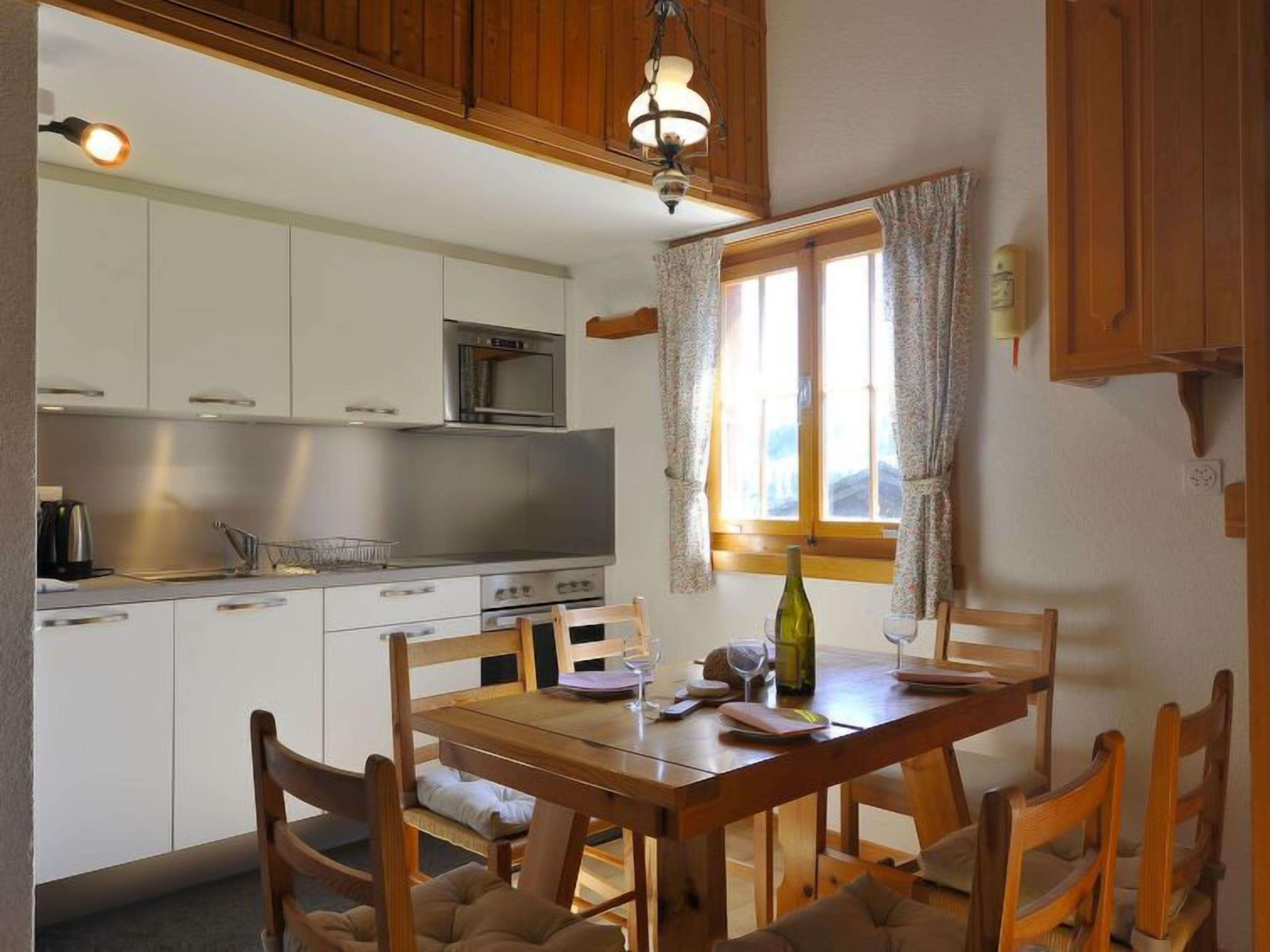 Une cuisine avec des meubles blancs, un four à micro-ondes et une table à manger en bois, accompagnée de chaises.