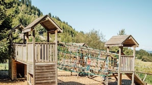 A wooden playground structure with a rope bridge and climbing net, set on a gravel surface with trees in the background. - Mountain View by Interhome (Moléson-sur-Gruyères)