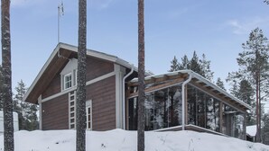 En stuga av trä med en glasveranda, omgiven av snötäckt mark och träd.