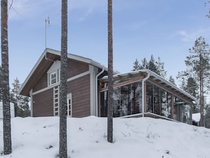 Une cabane en bois avec une extension en verre, entourée d’un paysage enneigé et d’arbres.