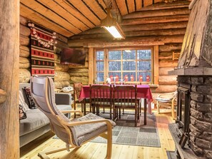 Acogedor interior de cabaña de troncos con comedor, chimenea, televisor y ventana con vistas a árboles.