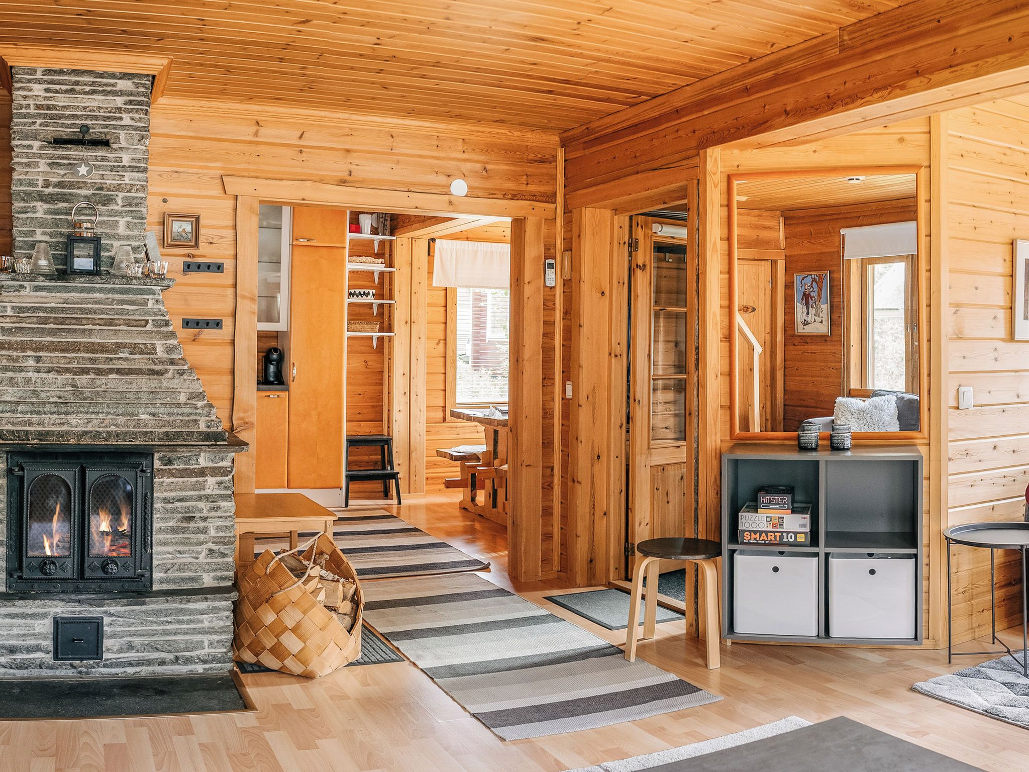 A cozy cabin interior with a stone fireplace, wooden walls, a dining area, and a kitchen with a refrigerator.