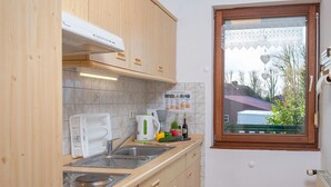 Een kleine keuken met houten kastjes, een fornuis, een spoelbak en een raam met uitzicht op een huis en bomen.
