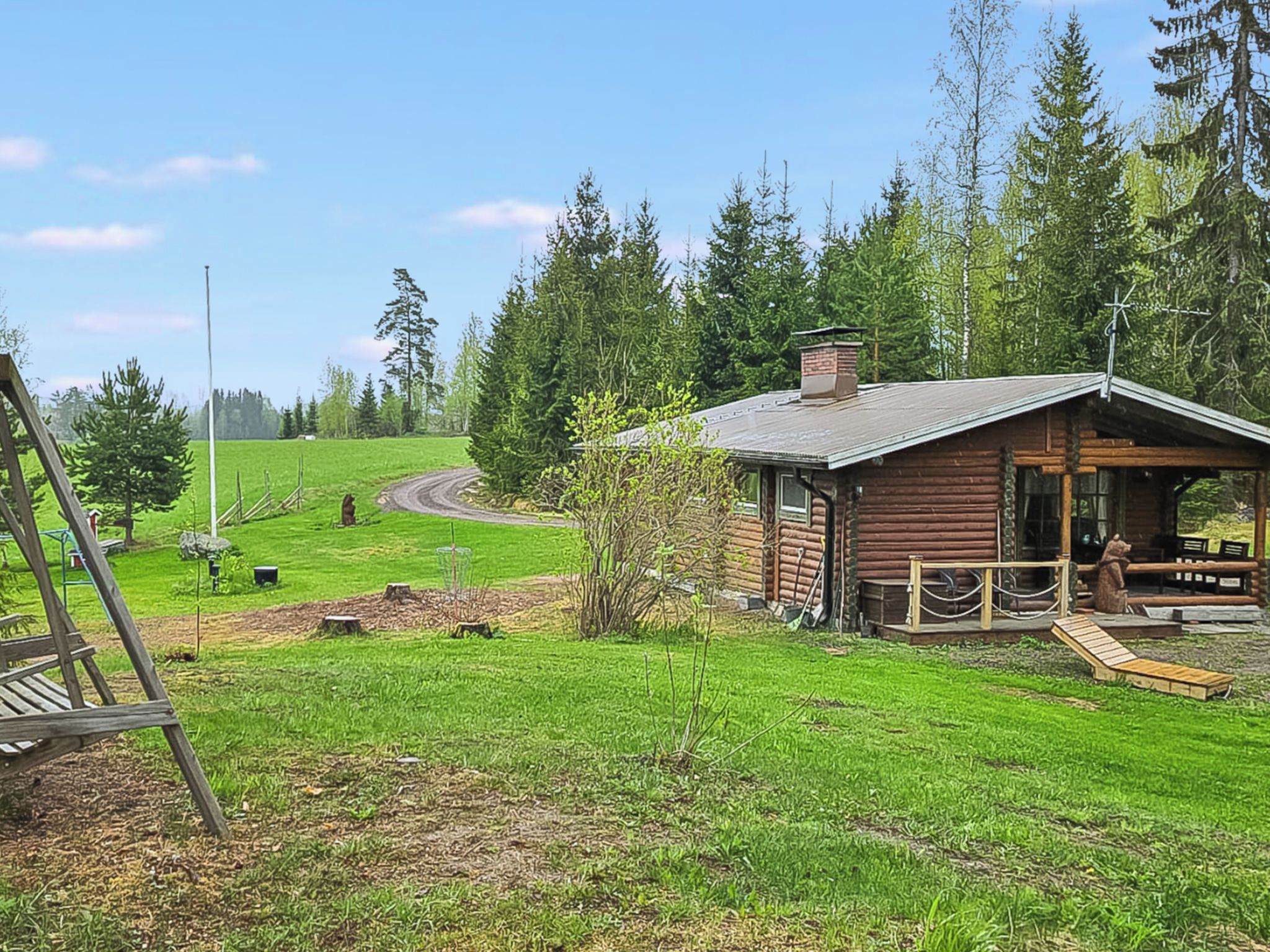 Tunnelmallinen mökki, jossa on puinen terassi ja katettu kuisti. Mökkiä ympäröi niitty ja tiheä metsä.