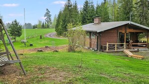 Una cabaña rústica con un porche, rodeada por un campo cubierto de hierba y un bosque denso.