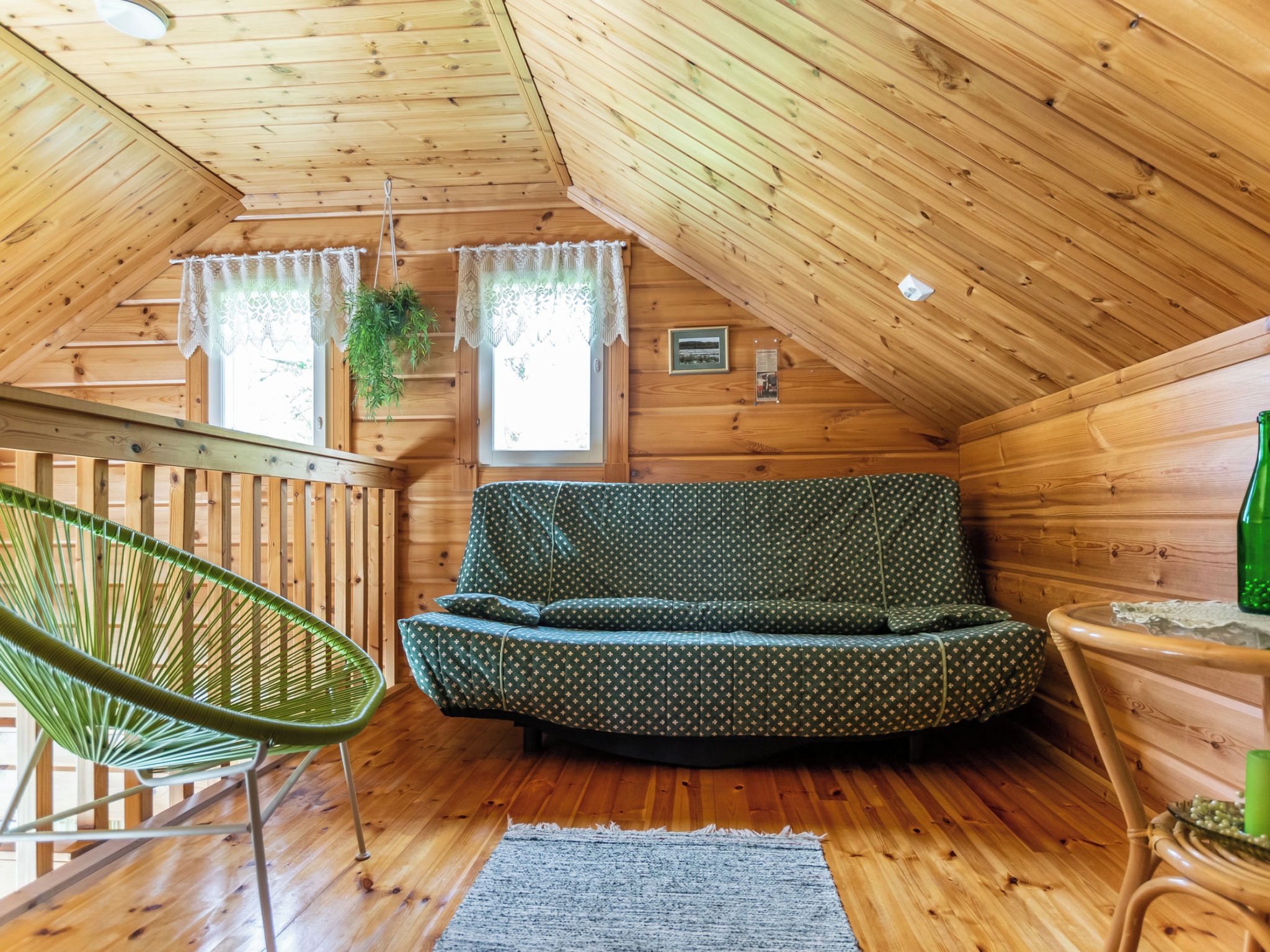 Una camera mansardata accogliente con soffitto spiovente in legno, un divano verde con motivi decorativi, una poltrona in vimini e un tavolino con una bottiglia.