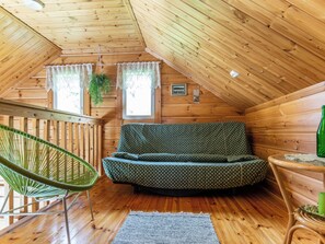 A cozy attic room with a wooden sloped ceiling, a green patterned sofa, a wicker chair, and a small table with a bottle.