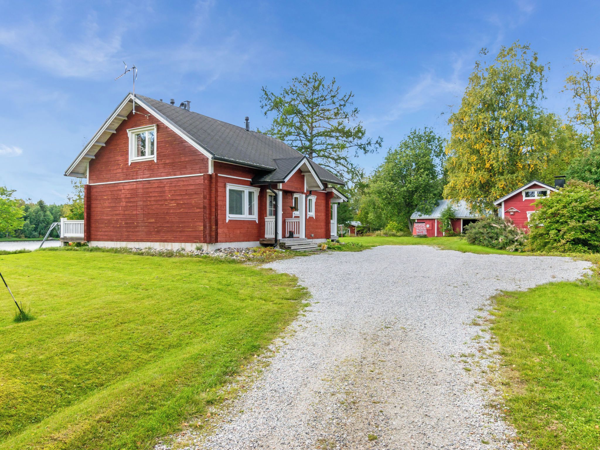 Een rode woning met een grindpad dat naar een witte deur leidt, omgeven door groen gras en bomen.