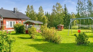 Een rode woning met een veranda, een schommel en een kleine tuinhuis in een groene tuin.