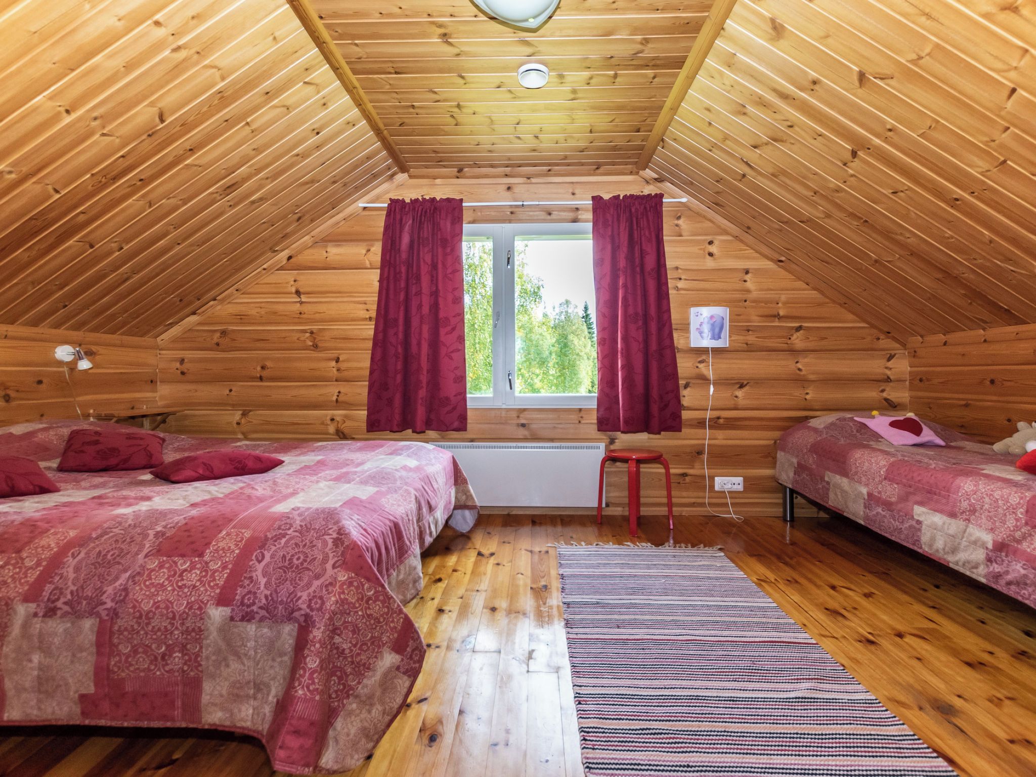 A cozy attic bedroom with two beds, a window with curtains, a radiator, and a small stool.