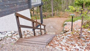 A wooden staircase leading to a white balcony with a metal railing, surrounded by a gravel path and trees. - Weekend duo by Interhome (Sotkamo)