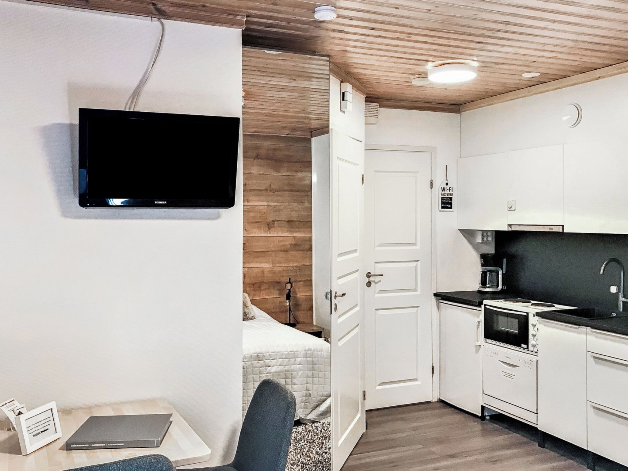 A compact hotel room with a flat-screen TV mounted on the wall, a kitchenette with a stove and microwave, and a bed with a white headboard.