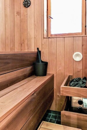 A wooden sauna room with benches, a black bucket, a white bowl, and a window.