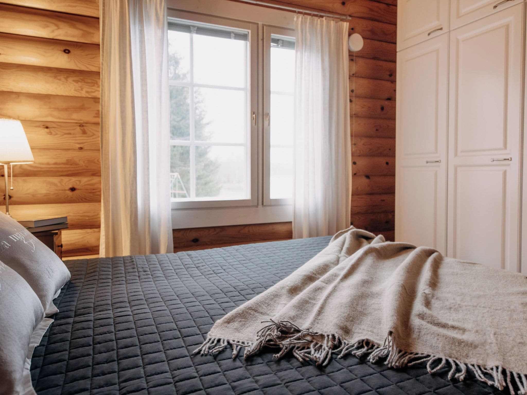 Un dormitorio con cama, ventana con cortinas, mesita de noche con lámpara y paredes de madera.