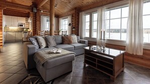 L’intérieur chaleureux d’un chalet en bois comprend un espace de vie avec un canapé, un pouf et une table basse, une cuisine dotée de meubles blancs et de grandes fenêtres ornées de rideaux.