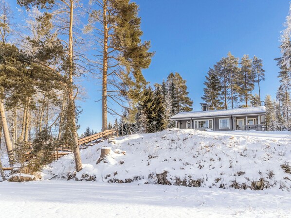 A cabin with a wooden deck and stairs leading up to it, surrounded by snow-covered ground and tall pine trees.