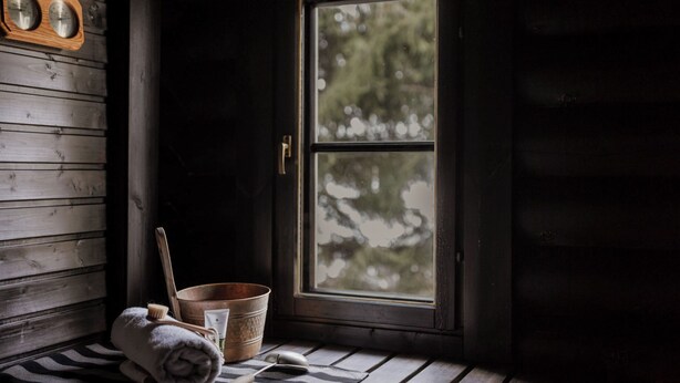 L’intérieur d’un chalet en bois, avec une fenêtre, un banc en bois, une serviette, un seau en métal et une horloge en bois.