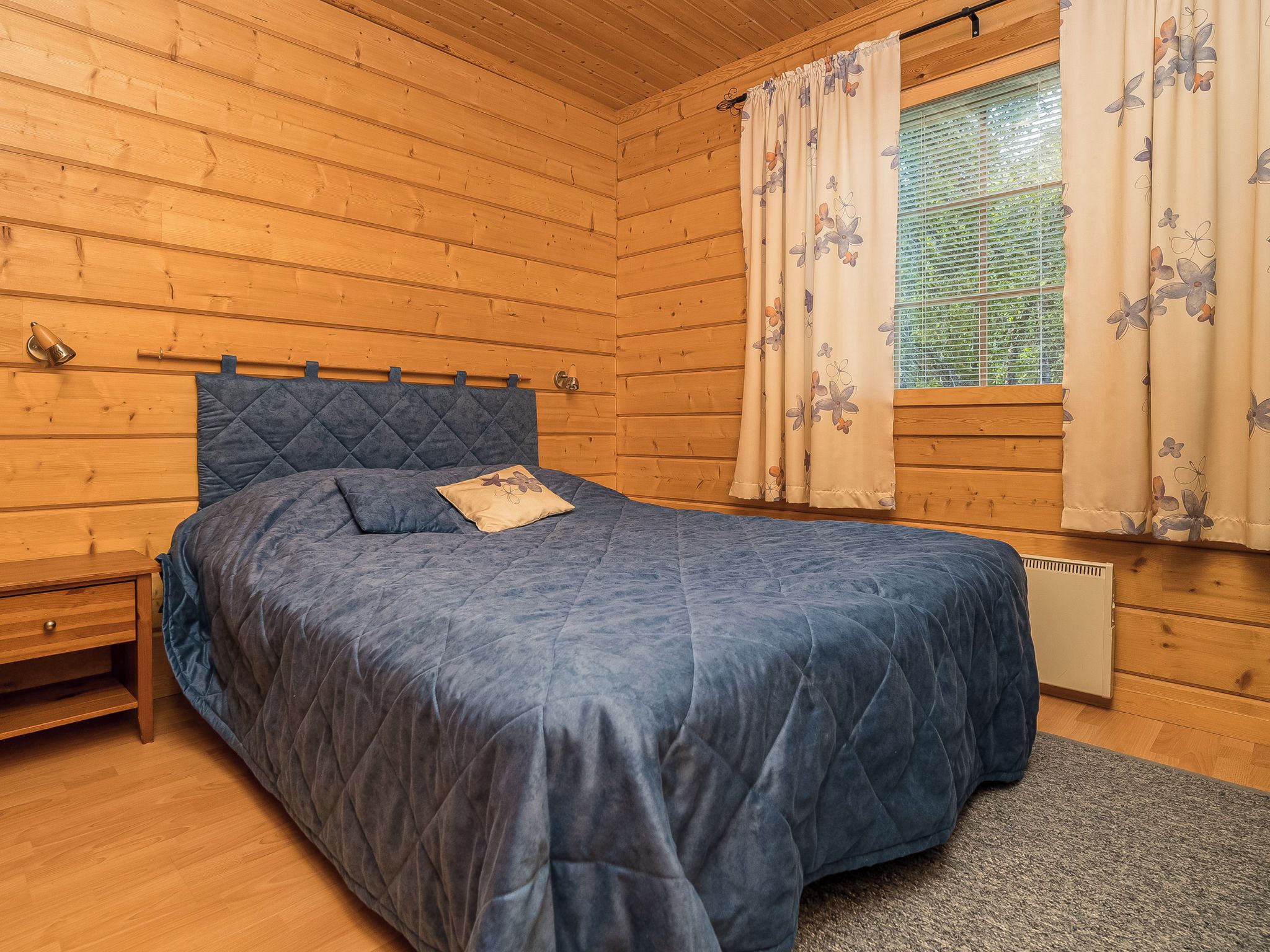 A wooden cabin bedroom with a bed, a nightstand, and a window with curtains.