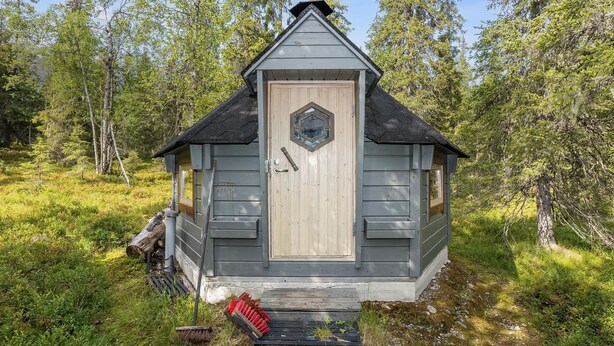 A small, single-story cabin with a wooden door and two windows, surrounded by a forested area.