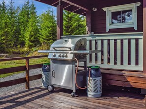 Ein tragbarer Grill auf einer Holzterrasse mit Geländer, zwei Müllbehälter und ein Fenster mit Blick auf einen Wald.