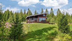Une cabane dotée d’une terrasse en bois et de grandes fenêtres, entourée d’une forêt dense et d’une zone herbeuse.