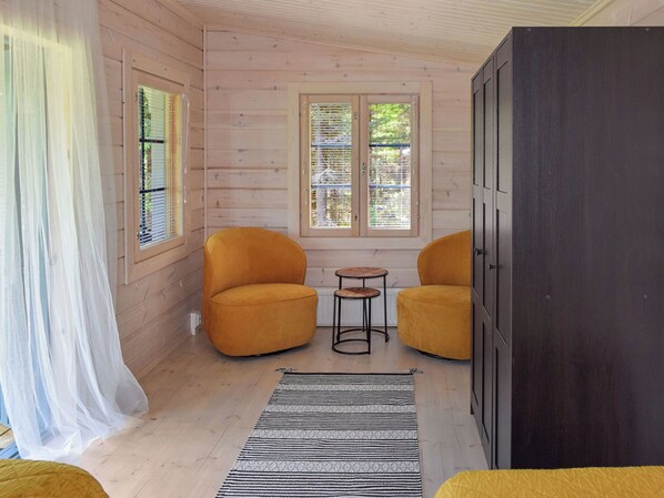 Una habitación con paredes de madera, dos sillas amarillas, una mesita redonda pequeña, un armario oscuro y un tapete a rayas.