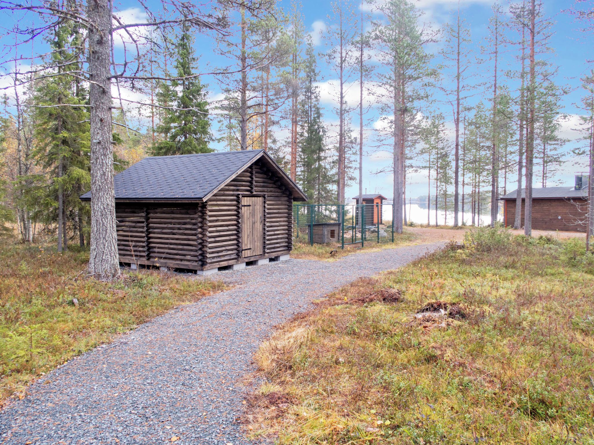 Une cabane en bois, accessible par un chemin de gravier, entourée de grands arbres et surplombée par un ciel dégagé.