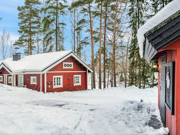 A red wooden cabin with a snow-covered roof, surrounded by tall pine trees.