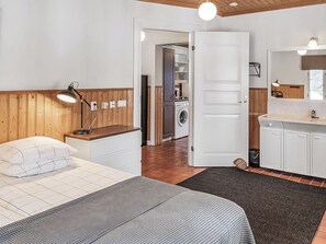 A bedroom with a bed, bedside table, lamp, and a view into the kitchen.