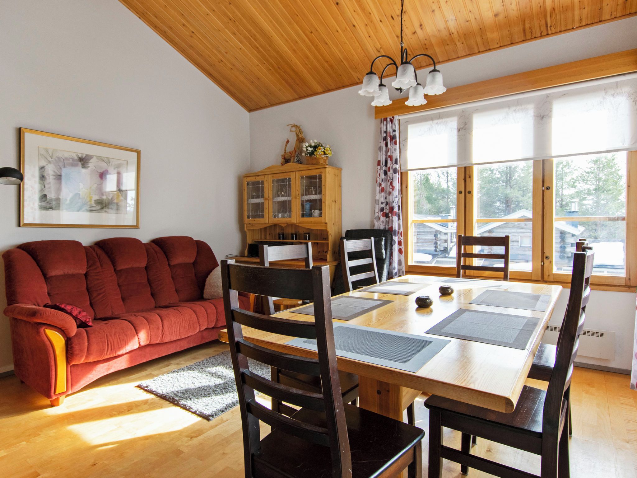 A cozy living room with a red sofa, wooden dining table, and chairs, a cabinet with glass doors, and a large window with a view of the outside.