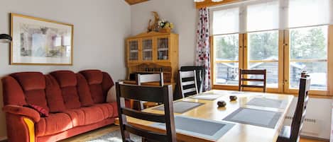 A cozy living room with a red sofa, wooden dining table, and chairs, a cabinet with glass doors, and a large window with a view of the outside.