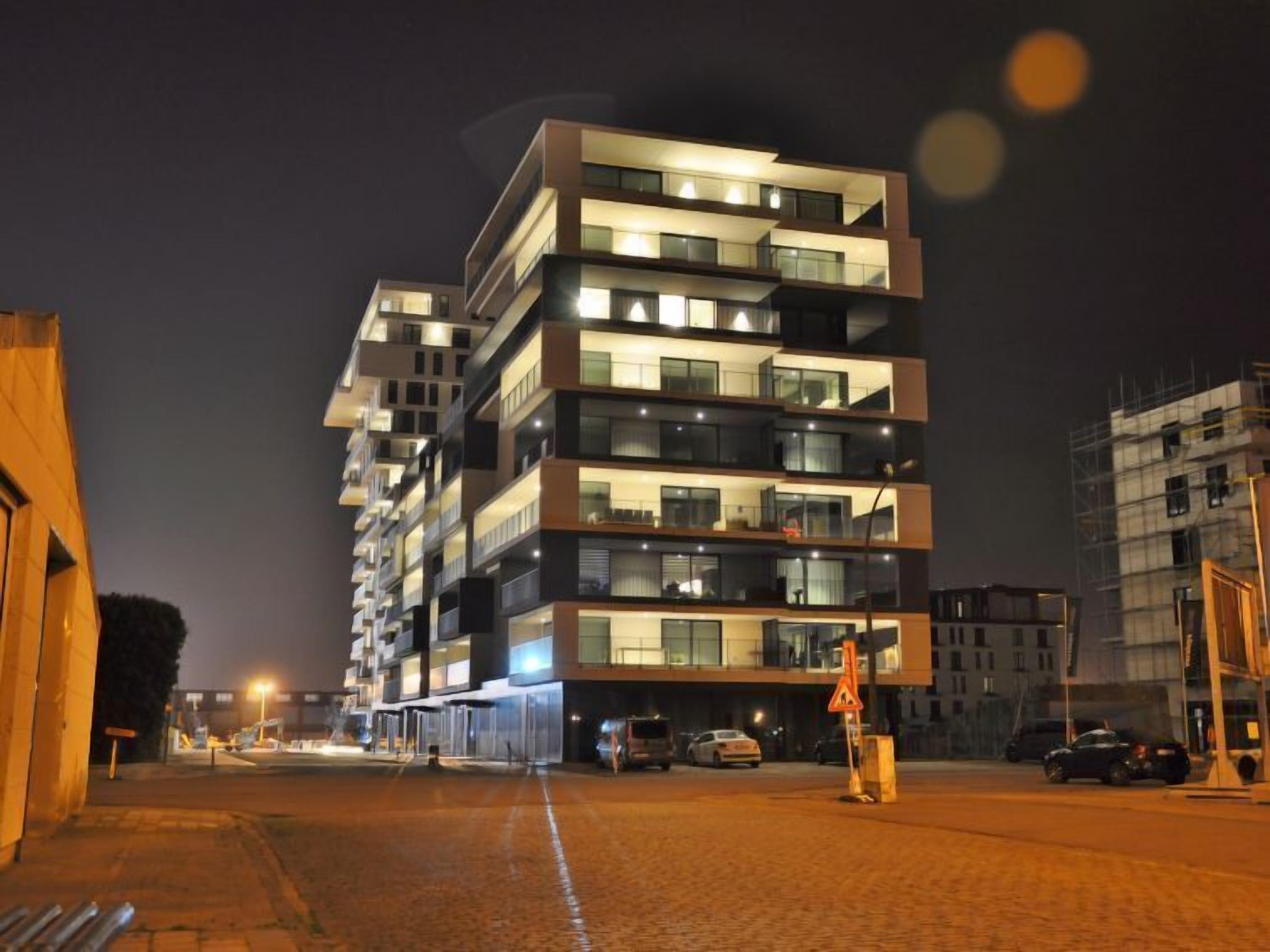 A multi-story hotel with balconies, a parking area, and a street with cars.