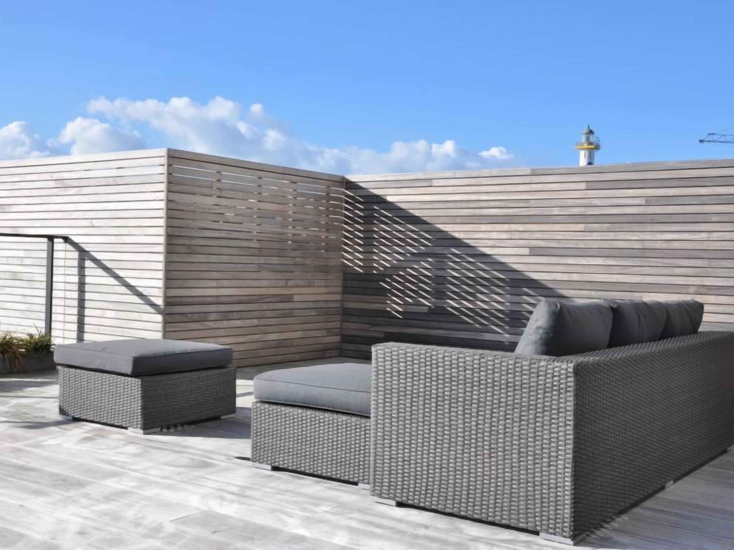 Un patio en la azotea con un área de estar moderna al aire libre, que incluye un sofá y dos otomanas, y una pared de madera con diseño de celosía.