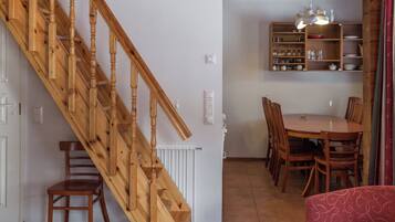 A wooden staircase leading to a dining area with a table and chairs, a red sofa, and a kitchen area with shelves.