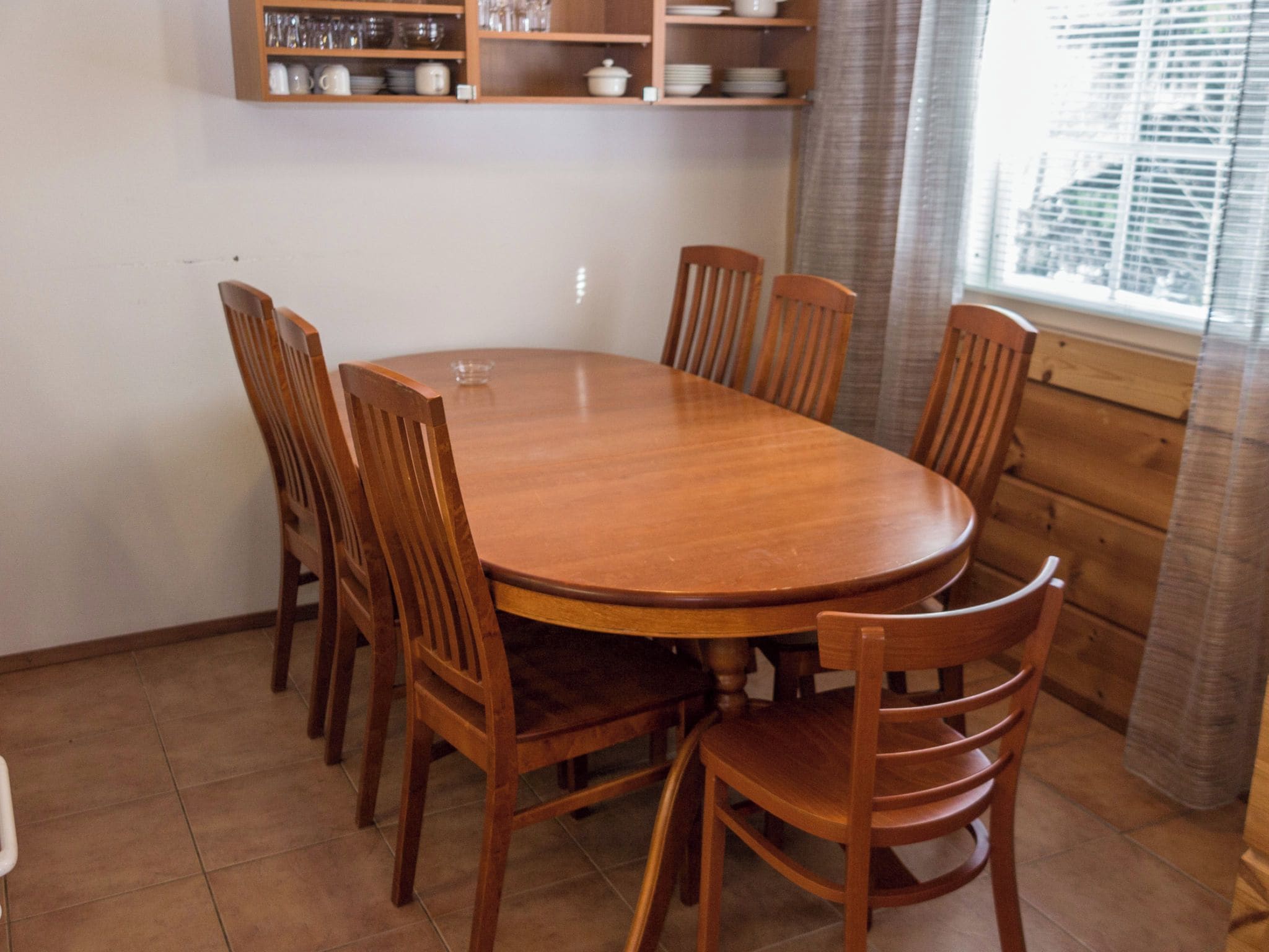 Una mesa de comedor redonda de madera rodeada por seis sillas, con una zona de cocina en lo alto y una ventana con cortinas a la derecha.
