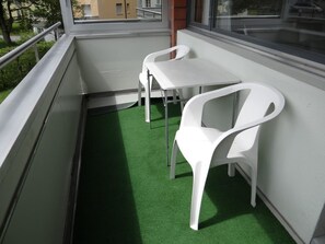 Un balcón con una pequeña mesa y dos sillas, piso de césped artificial verde y una ventana con vista a edificios.