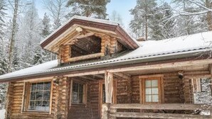 A rustic log cabin with a front porch and large windows, surrounded by snow-covered trees.