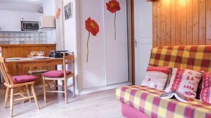 Une pièce chaleureuse avec un plancher en bois, un canapé à carreaux agrémenté de coussins roses, une table à manger en bois avec des chaises, et un coin cuisine en arrière-plan.